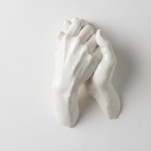Op de afbeelding: Een witte keramische sculptuur van twee in elkaar grijpende handen. De handen zijn glad en gedetailleerd, met zichtbare vingers en knokkels. De sculptuur is gemonteerd op een wit oppervlak, wat een minimalistische esthetiek cre&euml;ert.