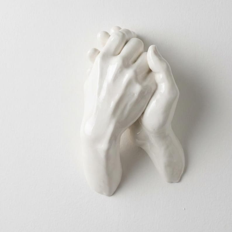 Op de afbeelding: Een witte keramische sculptuur van twee in elkaar grijpende handen. De handen zijn glad en gedetailleerd, met zichtbare vingers en knokkels. De sculptuur is gemonteerd op een wit oppervlak, wat een minimalistische esthetiek cre&euml;ert.