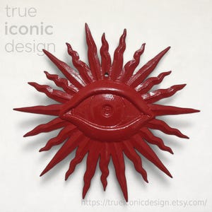May include: A bright red wall decoration featuring a central eye motif surrounded by radiating, wavy sunburst rays. The text 'true iconic design' appears in the upper left corner, and the URL 'trueiconicdesign.etsy.com' is at the bottom.