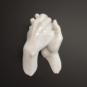 Op de afbeelding: Een witte keramische sculptuur van twee in elkaar grijpende handen. De handen zijn glad en glanzend, met zichtbare details van vingers en knokkels. De sculptuur staat tegen een donkere achtergrond.