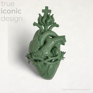 Könnte beinhalten: Eine salbeigrüne Herzskulptur mit anatomischen Details, einer Dornenkrone und einem Kreuz. Die Worte "true iconic design" befinden sich in der oberen linken Ecke. Die Skulptur hat eine glänzende Oberfläche.
