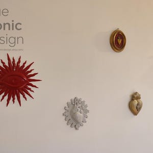 May include: A collection of wall-mounted decorative objects including a red sunburst eye, a small oval framed heart, a large yellow anatomical heart, a silver anatomical heart with rays, and a gold heart. Text reads true iconic design.