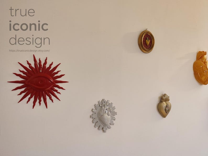 May include: A collection of wall-mounted decorative objects including a red sunburst eye, a small oval framed heart, a large yellow anatomical heart, a silver anatomical heart with rays, and a gold heart. Text reads true iconic design.