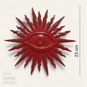 May include: A bright red wall ornament shaped like a sun with a central eye, featuring wavy rays extending outward. The object measures 23 cm in height as indicated by the vertical line and text. The text 'true iconic design' is visible below.