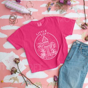 May include: Pink t-shirt with a white graphic design of a house with the text "Little Loft" and "Mocks" on a pink and white background.