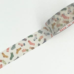 以下が含まれることがあります： さまざまなデザインのカラフルな靴下の繰り返し模様が特徴の和紙テープロール。 靴下は、クリーム色の背景にピンク、緑、黄色、青の色合いです。 テープには「MADE IN JA」と表示されています。