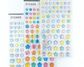 Puede incluir: Tres hojas verticales de pegatinas en envases de plástico transparente. Las hojas presentan filas de pequeñas pegatinas en relieve, brillantes y de colores pastel con formas de corazón, flor y estrella. Cada encabezado lleva la palabra STICKER.