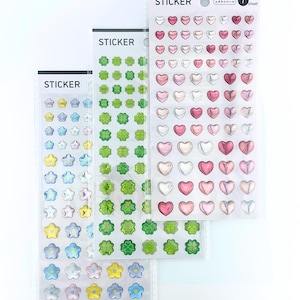 May include: Three vertical sheets of stickers labeled STICKER at the top. One sheet features pink and white heart-shaped stickers, one features green four-leaf clover stickers, and one features blue and yellow flower-shaped stickers.