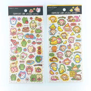 Puede incluir: Dos hojas de pegatinas de la serie TOPPING LAB hechas en Japón. La hoja izquierda presenta gatos, ranas y fresas con texto que incluye 'HELLO' y 'TOMATO'. La hoja derecha muestra monos y plátanos con texto que incluye 'monkey' y 'BANANA'.