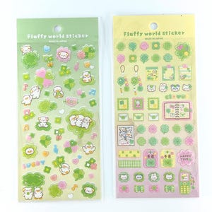 May include: Two sheets of Fluffy world sticker sets made in Japan. The stickers feature green clovers, small white animals, and pink floral accents. Text on the sheets includes "Fluffy world sticker," "LUCKY," "HAPPY," "clover," and "HAPPY TIME."