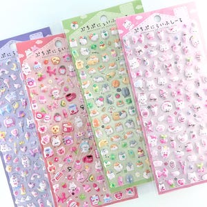 以下が含まれることがあります： 紫、ピンク、緑、薄ピンクの4枚のぷっくりとしたシールシート。各シートには動物、食べ物、リボン、星などの小さな立体的なアイコンが並んでおり、上部には「ぷちぷにらいふ」という日本語のテキストが印刷されています。
