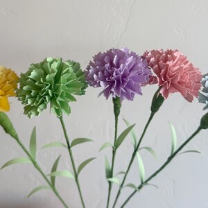 May include: Five paper carnations in different colors: yellow, green, purple, pink, and blue. The carnations are arranged in a bouquet with green stems.