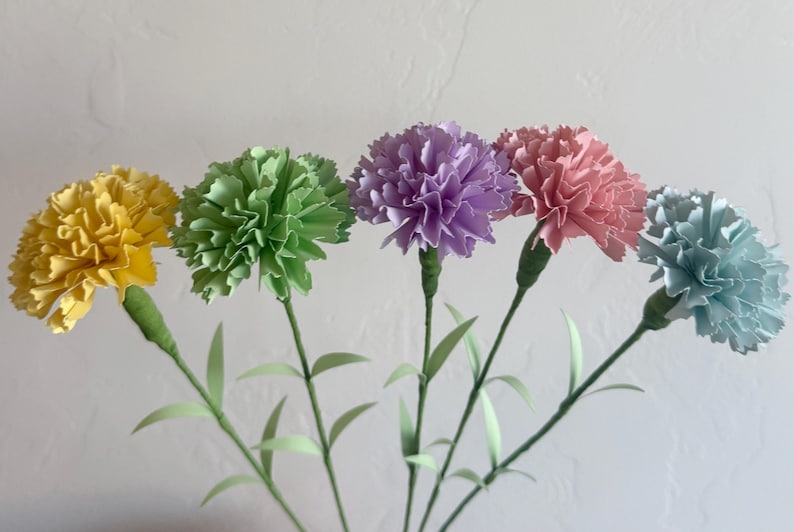 May include: Five paper carnations in different colors: yellow, green, purple, pink, and blue. The carnations are arranged in a bouquet with green stems.