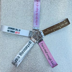 May include: Five fabric keychains arranged in a star pattern, each with a metal ring and clasp. The straps feature text: 'to live for the hope of it all', 'you're a cowboy like me', 'I'm a real tough kid', 'getaway car', and 'A LOT GOING ON AT THE MOMENT'.