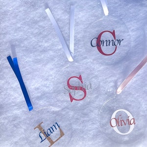 May include: Four clear round ornaments with different names and initial letters written in glitter. The names are Connor, Sophia, Liam, and Olivia. Each ornament has a ribbon attached.