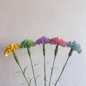 May include: Five paper carnations in different colors: yellow, green, purple, pink, and light blue. The carnations are arranged in a row with their stems pointing downwards. The carnations are against a white background.