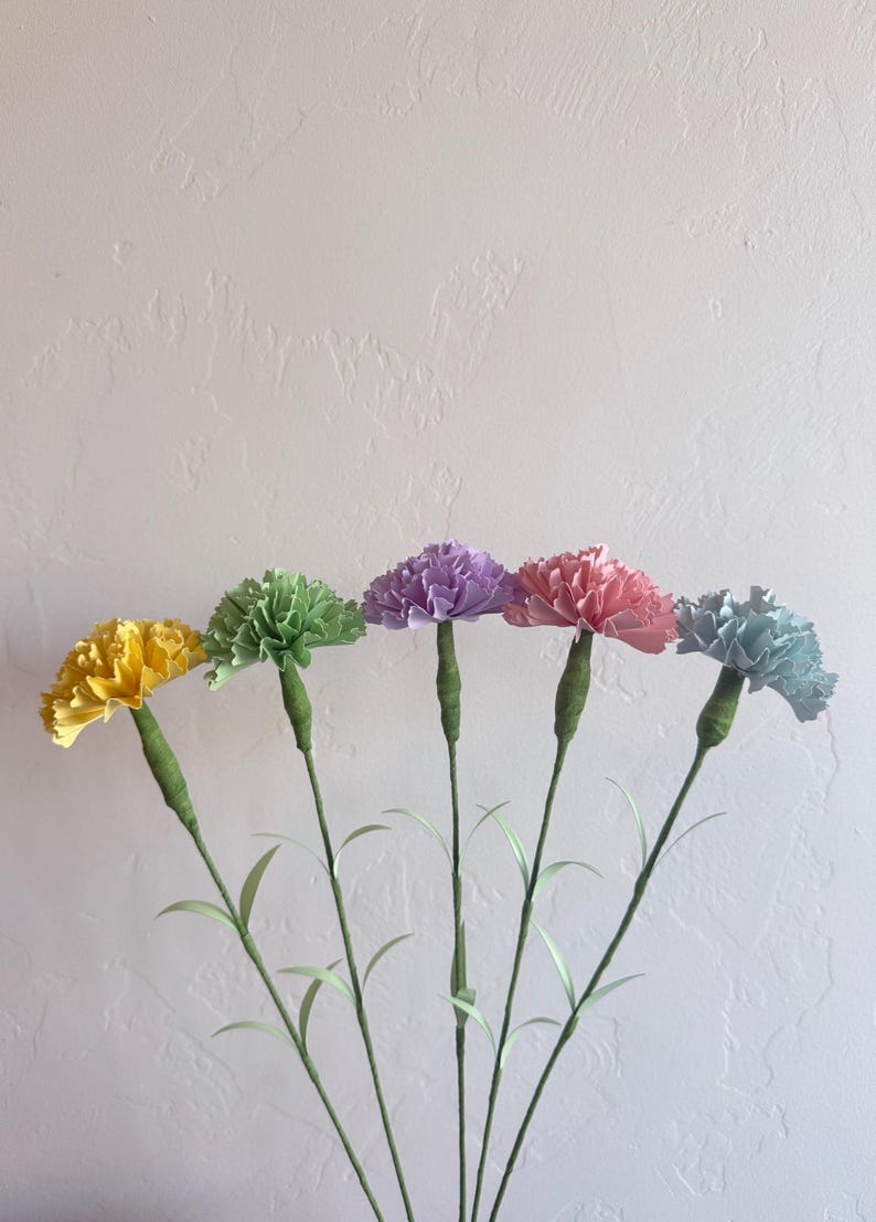 May include: Five paper carnations in different colors: yellow, green, purple, pink, and light blue. The carnations are arranged in a row with their stems pointing downwards. The carnations are against a white background.