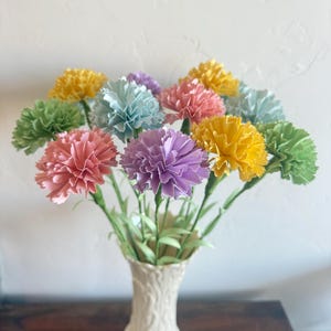 May include: A bouquet of pastel paper carnations in a white ceramic vase. The carnations are in shades of yellow, green, pink, blue, and purple.