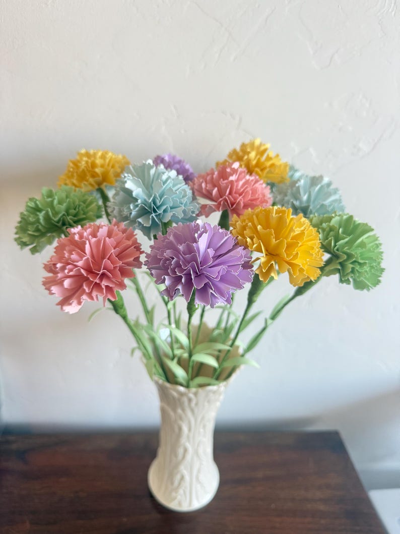 May include: A bouquet of pastel paper carnations in a white ceramic vase. The carnations are in shades of yellow, green, pink, blue, and purple.