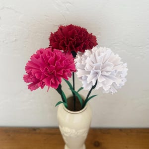 May include: A cream-colored vase holds three artificial carnations. One is deep red, another is bright pink, and the third is white. The flowers have green stems and leaves. The vase sits on a wooden surface.