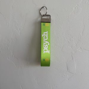 May include: A green keychain with the word "Psych" in white lettering. The keychain has a silver metal clasp and key ring. The green strap has a repeating pattern of yellow pineapples.