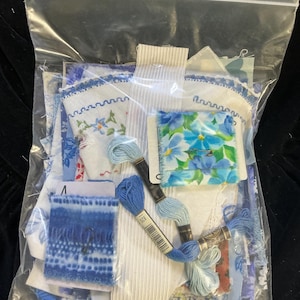 May include: A clear plastic bag filled with blue and white fabric scraps, embroidery floss, and a floral pin. The fabric pieces feature floral patterns and lace trim. Several spools of blue embroidery floss are visible.