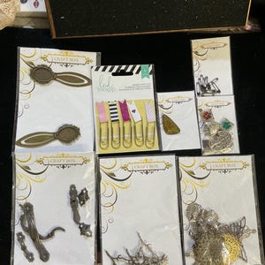 May include: A collection of craft supplies, including metal embellishments, paper clips, and decorative accents. The items are packaged on white cards with gold accents and the "Craft Box" logo. Various shapes and designs are visible, including floral and ornate metal pieces.