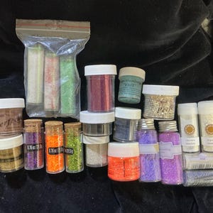 May include: A collection of various glitter containers and vials. The assortment includes jars of gold, purple, orange, and green glitter, along with clear vials of white and iridescent glitter. Some containers have labels with text.