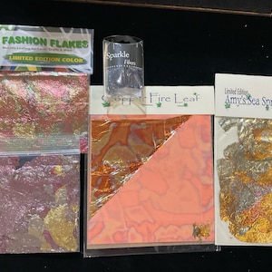May include: Four packages of metallic leaf flakes for crafts. The packages are labeled "Fashion Flakes Limited Edition Color", "Sparkle Fibers", "Copper Fire Leaf", and "Amy's Sea Spray Leaf Limited Edition by Amy's Magic".