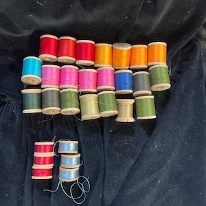 May include: A collection of sewing thread spools in various colors including red, orange, yellow, pink, blue, green, gray, and tan. The spools are made of wood and are arranged in a grid-like pattern, with some smaller spools positioned separately.