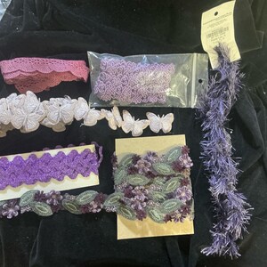 May include: Assortment of craft supplies including various ribbons, lace, and embellishments. The items are in shades of purple, lavender, and white. There are butterfly and floral designs, and a length of fuzzy trim.