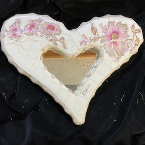 May include: Heart-shaped mosaic mirror with a central opening. The white mosaic frame is adorned with pink floral designs. The mirror is a decorative piece, suitable for wall decor.