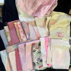 May include: A collection of assorted fabric scraps in various shades of pink, cream, and yellow. The pieces feature diverse textures, including smooth silk, textured crepe, and patterned weaves with floral and geometric motifs in multiple sizes.