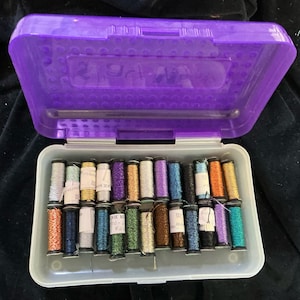 May include: An open purple plastic container filled with colorful spools of thread. The thread colors include silver, gold, blue, purple, orange, and green. The container is on a dark background, suggesting a sewing or craft project.