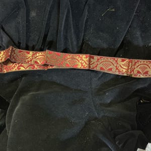 May include: A decorative ribbon with a red and gold floral pattern. The ribbon is lying on a black velvet surface. The ribbon's design features intricate details and a contrasting color scheme.