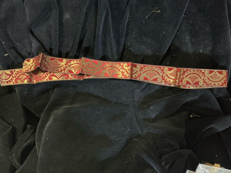 May include: A decorative ribbon with a red and gold floral pattern. The ribbon is lying on a black velvet surface. The ribbon's design features intricate details and a contrasting color scheme.