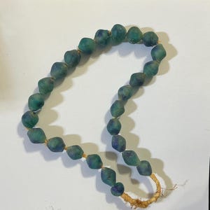 May include: A necklace featuring a strand of rounded, teal and blue beads. The beads are strung together with a natural fiber cord. The necklace is displayed on a white surface.