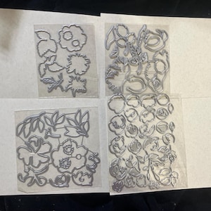 May include: Four sets of metal floral die cuts in various shapes and sizes, including flowers, leaves, and stems. The die cuts are silver and arranged on white paper.
