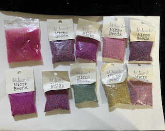 May include: A collection of small plastic bags filled with colorful micro beeds. The bags are labeled "Art Accents Micro Beedz" and contain various colors, including pink, purple, green, and gold. These craft supplies are ideal for jewelry making and other DIY projects.