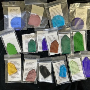 May include: A collection of small, colorful glass pieces in various shapes, including circles and tags, each sealed in a clear plastic bag. Each bag contains a printed label with text. The glass pieces are in shades of blue, green, purple, yellow, and red.