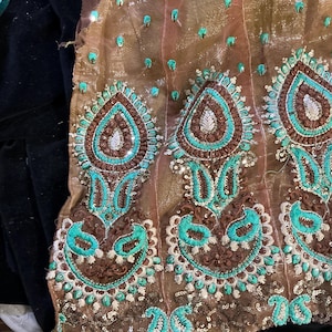 May include: Close-up of a section of brown fabric with intricate embroidery. The design features large, teardrop-shaped motifs in teal and brown, embellished with sequins. The fabric has a shimmering quality, suggesting it may be used for formal wear or decorative purposes.