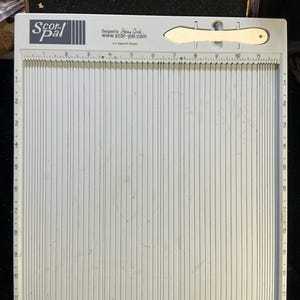 May include: A white Scor-Pal scoring board with vertical score lines and measurements. The board includes a bone folder tool and text that reads "Scor-Pal" and "Designed by Stina Crick www.scor-pal.com".