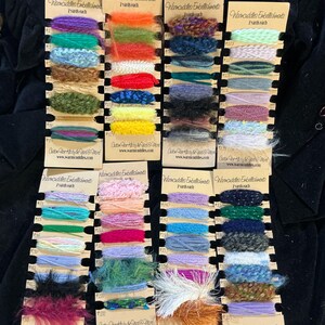 May include: A collection of yarn samples in various colors, including teal, pink, and purple, displayed on small cardboard tags. Each tag is labeled with "Warmcuddles Embellishments" and the website address. The yarn appears to be for crafting or embellishment purposes.