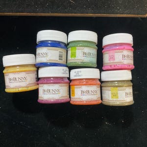 May include: An assortment of small jars of BoBunny glitter paste in various colors, including gold, pink, orange, green, and blue. Each jar has a white lid and a label with the brand name. The jars are arranged on a dark surface.
