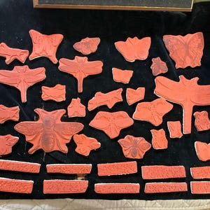 May include: A collection of red rubber stamps featuring various insect designs, including butterflies, dragonflies, and bees. The stamps are arranged on a black surface and include several rectangular stamps with a repeating pattern.