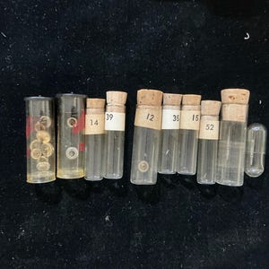 May include: A collection of small glass vials with cork stoppers, some containing tiny watch parts. Each vial has a label with a number. The vials are arranged on a dark surface.