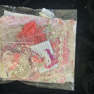 May include: A clear plastic bag containing a collection of craft supplies, including various pieces of floral and patterned fabric, lace trim, and small bundles of embroidery thread in shades of pink, red, white, and beige, with an instructional card.
