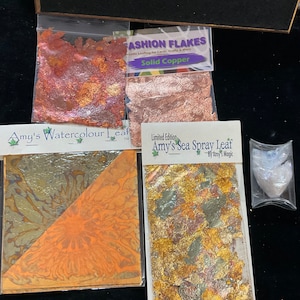 May include: A close-up of three packages of metallic leafing for crafts. The packages are labeled "Fashion Flakes Solid Copper", "Amy's Watercolour Leaf", and "Limited Edition Amy's Sea Spray Leaf By Amy's Magic".