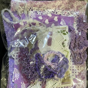 May include: A collection of purple fabric scraps, lace, and embroidery floss in a clear plastic bag. The fabrics feature floral, heart, and polka dot patterns. Various shades of purple and white are visible, with a few pieces of lace and a bundle of embroidery floss.