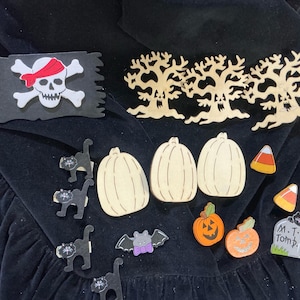 May include: A collection of Halloween-themed wooden cutouts including a black pirate flag with a skull, three spooky trees, three plain pumpkins, four black cats, a bat, two candy corns, two jack-o'-lanterns, and a tombstone labeled M.T. Tomb.